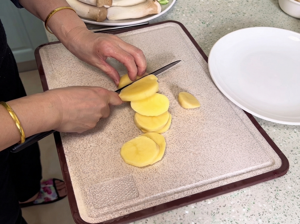 Patatas siendo cortadas con un cuchillo en una tabla de cortar al lado de un plato blanco.