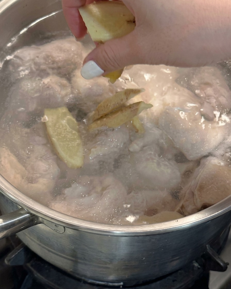 Una mano dejando caer rodajas de jengibre fresco en una olla de agua hirviendo con pollo.