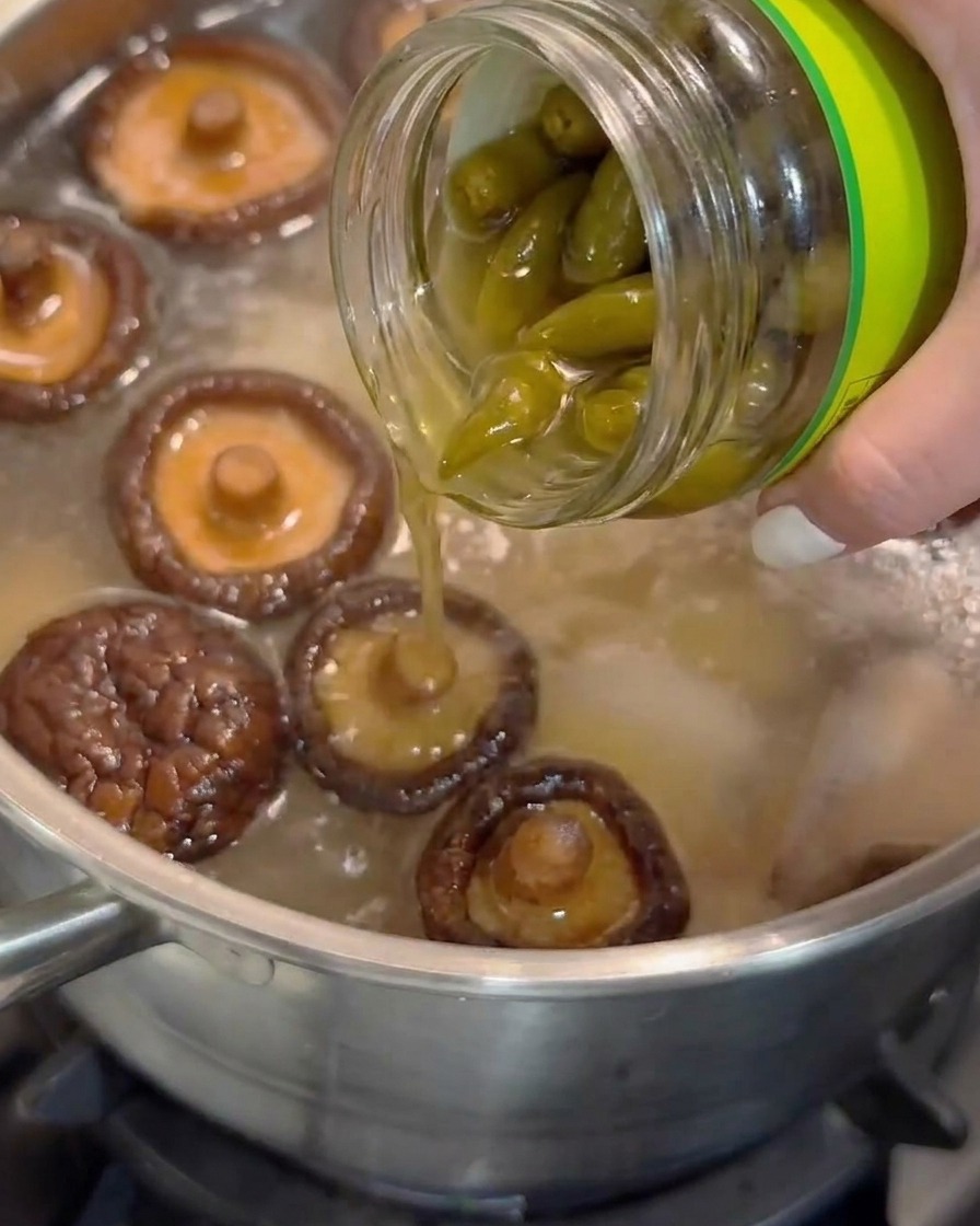 Jugo de chiles pelados en vinagre vertiéndose de un frasco de vidrio en una olla de sopa a fuego lento con setas shiitake.