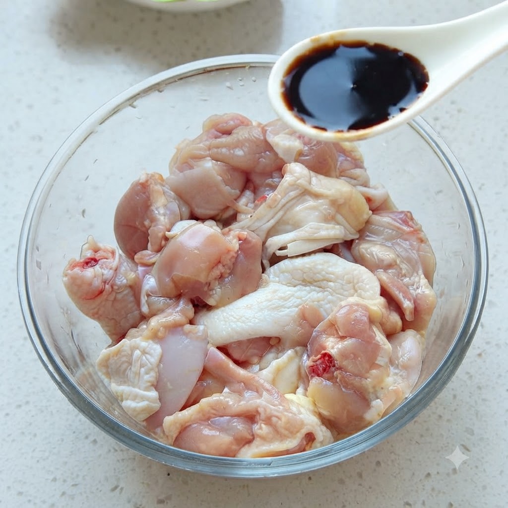 Una cuchara de salsa de soja oscura siendo añadida a un bol de vidrio lleno de trozos de pollo crudo.
