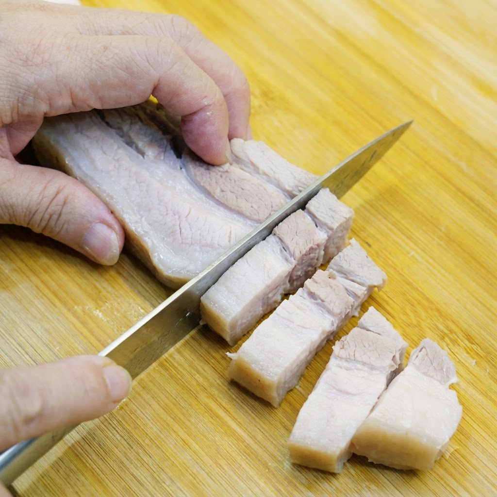 Panceta de cerdo blanqueada siendo cortada en trozos tamaño bocado en una tabla de madera.