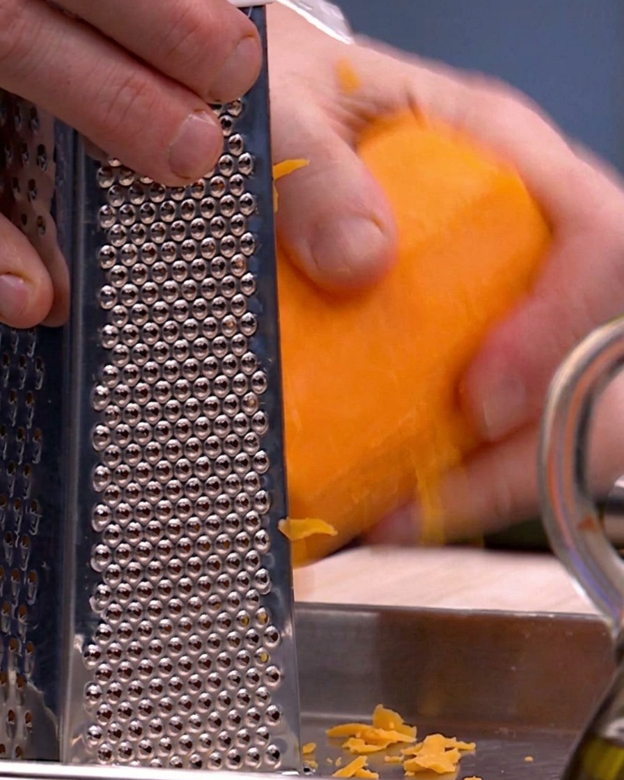 Manos usando un rallador de caja de metal para rallar calabaza butternut de color naranja brillante sobre una bandeja de metal.