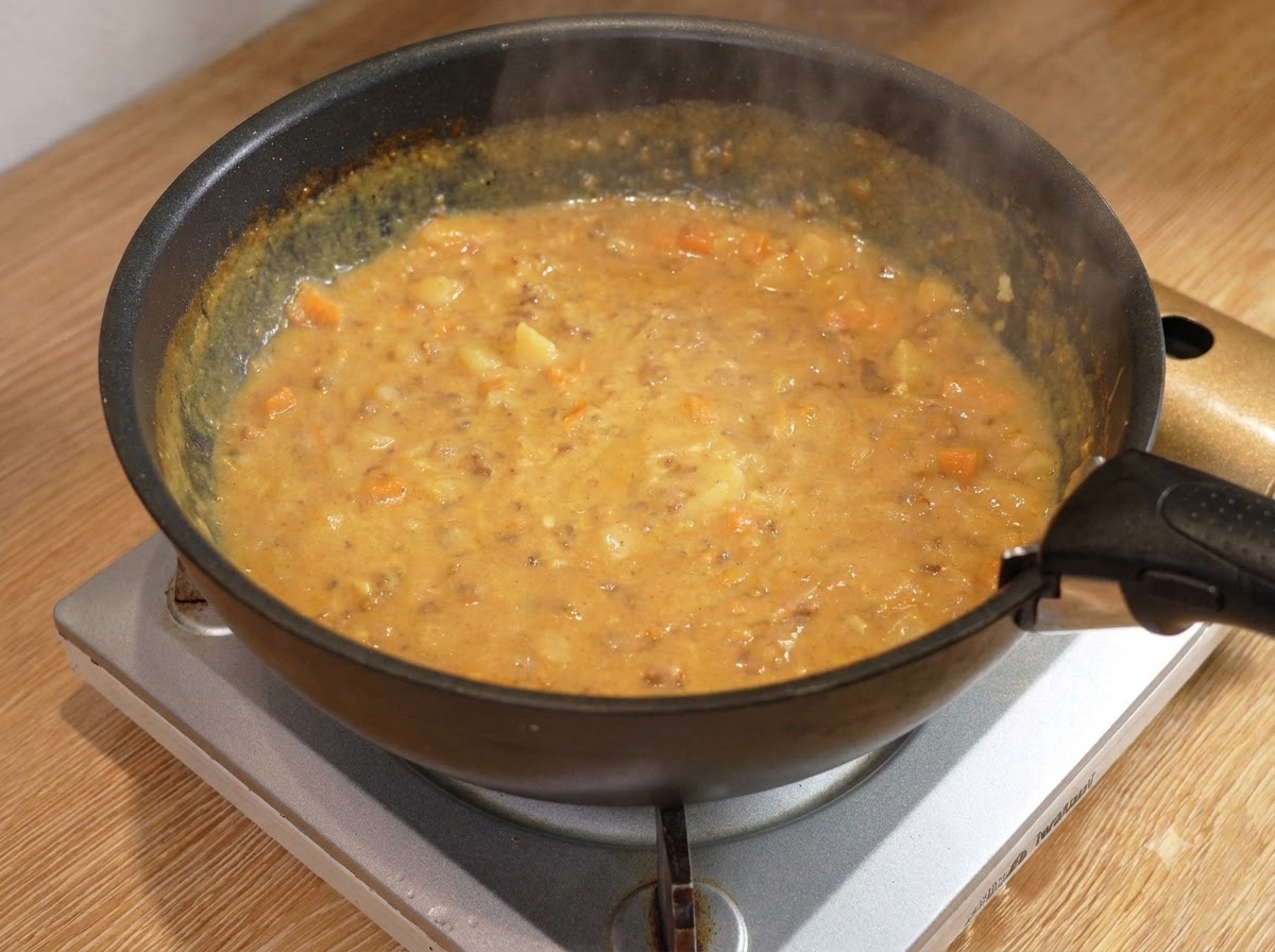 Una cuchara de madera removiendo una salsa de curry con carne espesa y burbujeante dentro de una sartén negra en un quemador.