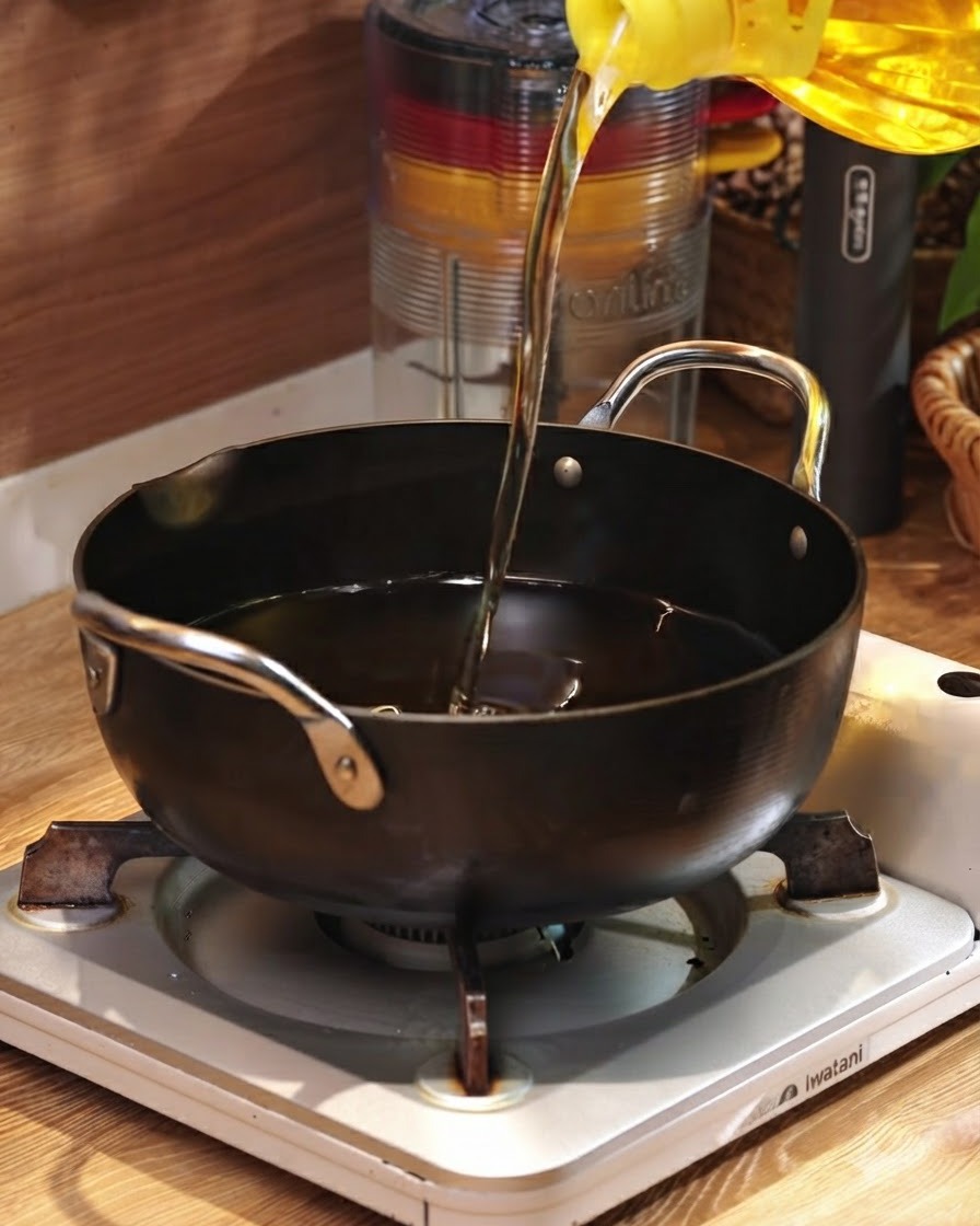 Aceite de cocina vertiéndose desde un recipiente amarillo en una sartén negra apoyada en un quemador portátil.