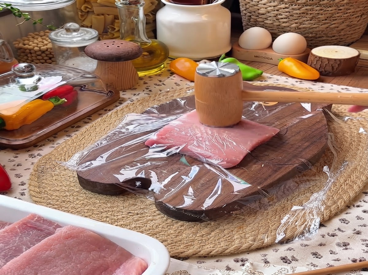 Un mazo de carne de madera golpeando un trozo de lomo de cerdo cubierto con film transparente en una tabla de cortar de madera.