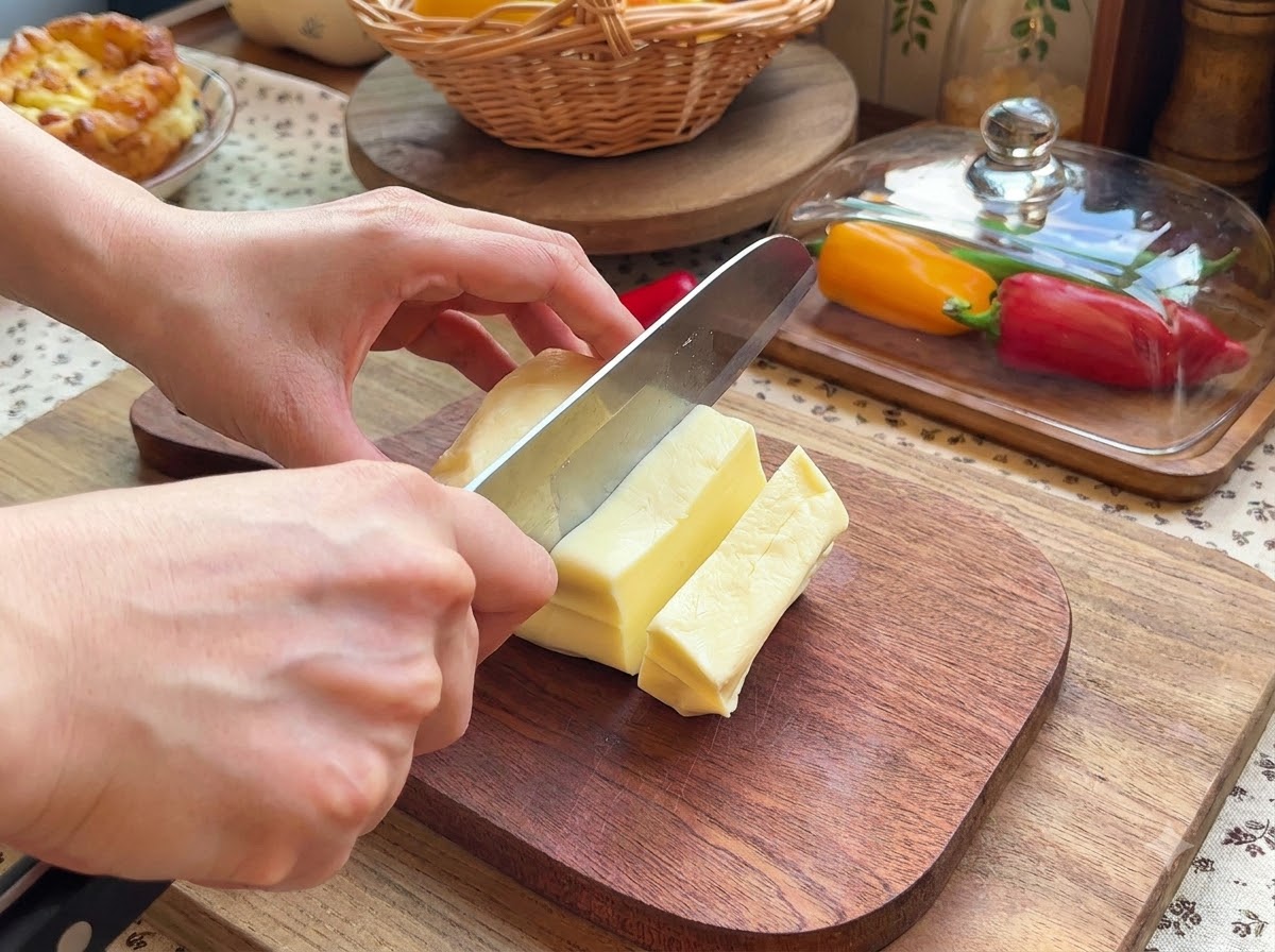 Un par de manos usando un cuchillo para rebanar un bloque de queso mozzarella en trozos rectangulares pequeños sobre una tabla de cortar de madera.