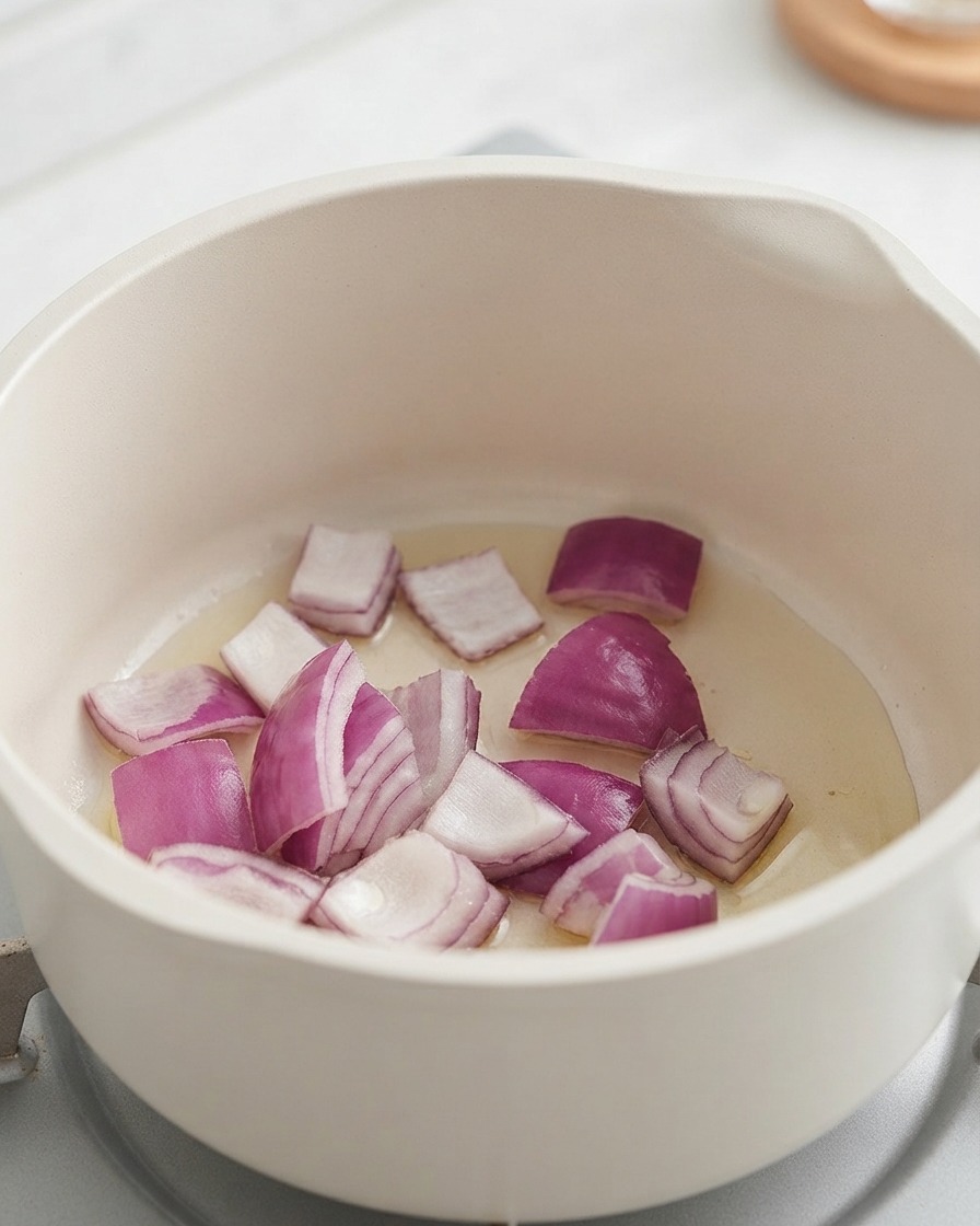 Cebollas rojas picadas y patatas cocinándose dentro de una olla blanca.