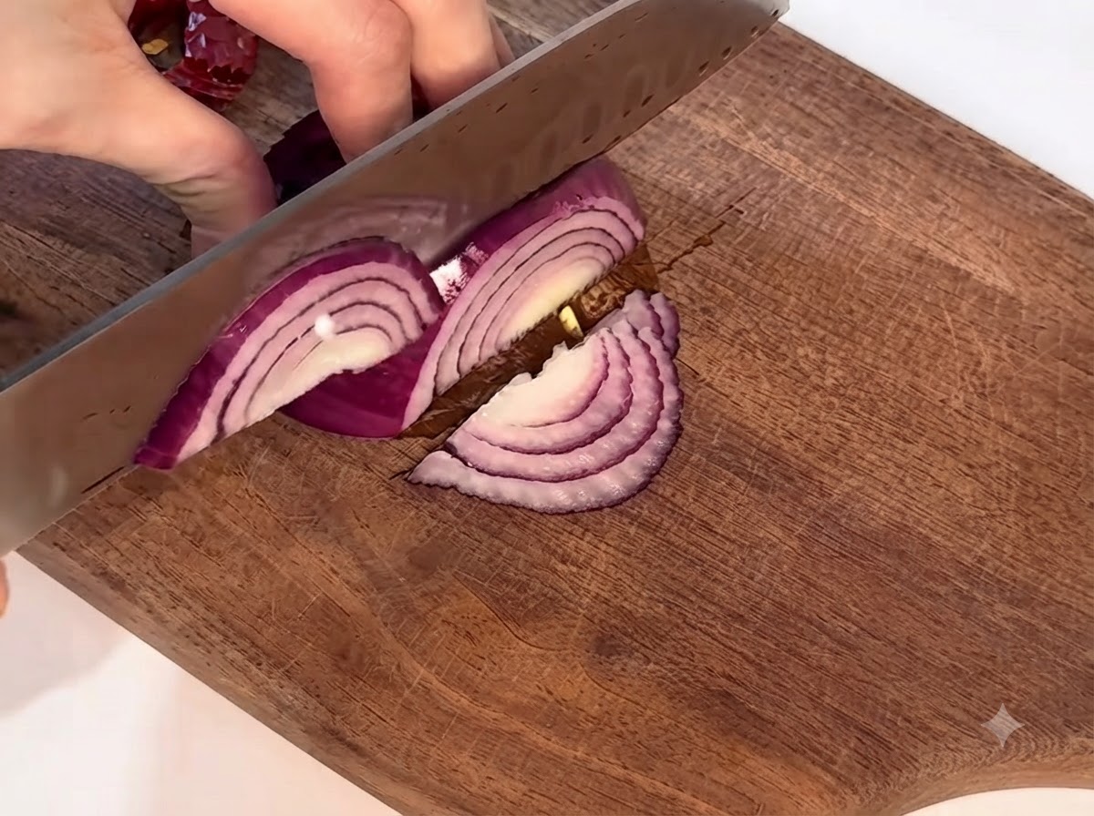Una cebolla morada siendo cortada cuidadosamente en una tabla de cortar de madera con un cuchillo.