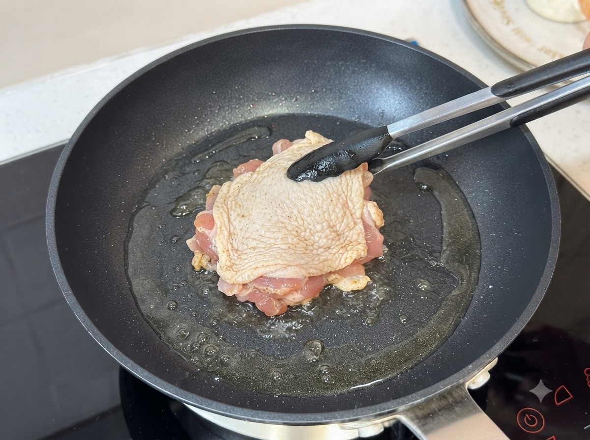 Un muslo de pollo sazonado friéndose con la piel hacia abajo en una sartén caliente con aceite usando pinzas.
