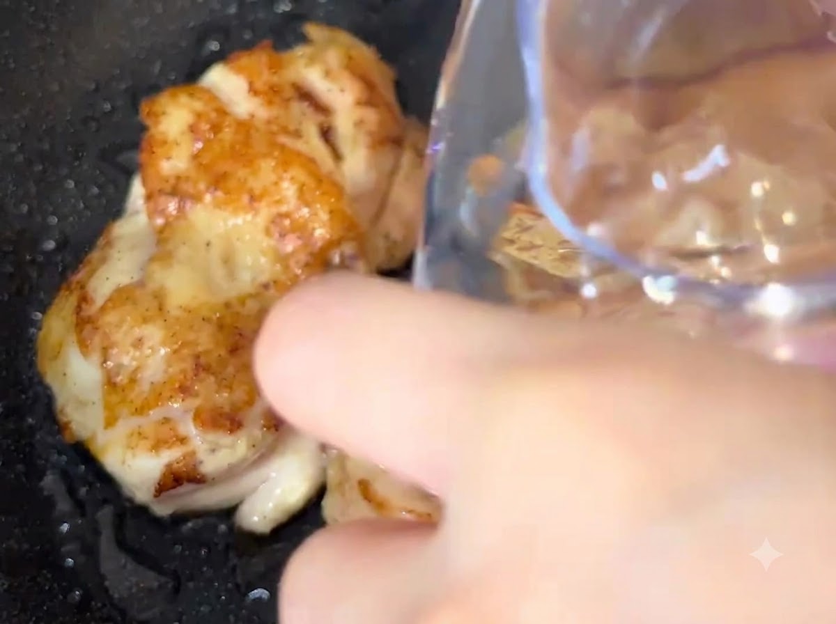 Agua siendo vertida de un envase directamente en una sartén con trozos de pollo fritándose.