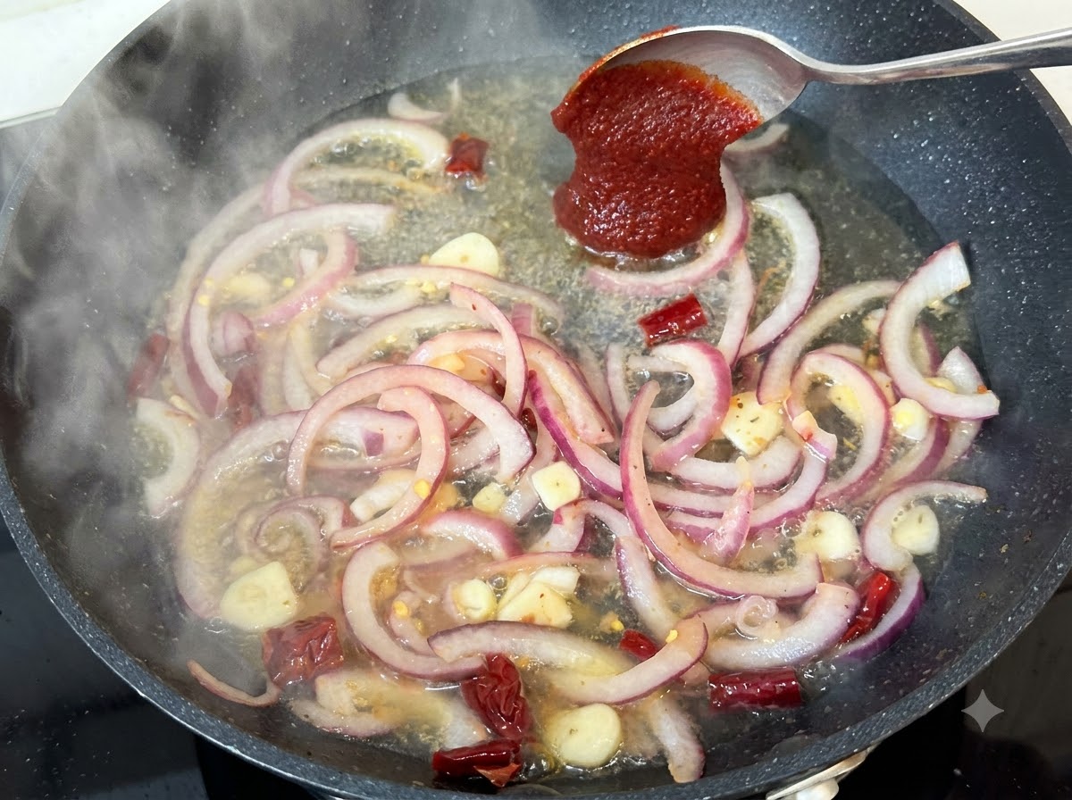 Una cucharada de pasta de chile coreano siendo añadida al líquido hirviendo a fuego lento de la sartén con cebollas y especias.