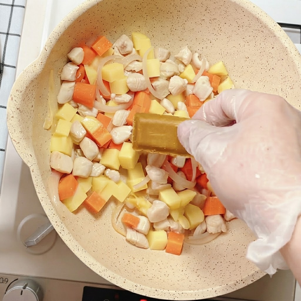 Una mano colocando un bloque de condimento de curry en la sartén llena de verduras mixtas y pollo.