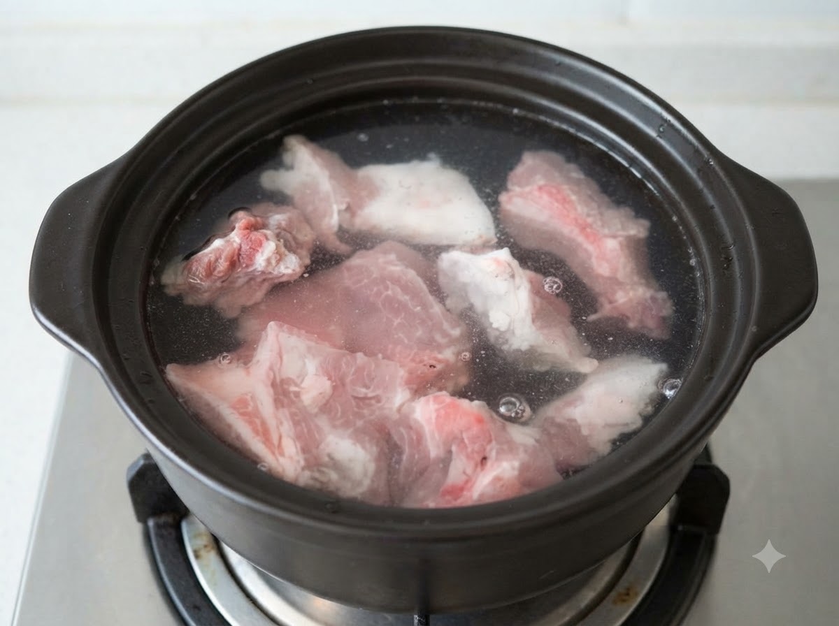 Trozos de hueso de cerdo crudos sumergidos en agua fría dentro de una olla de barro oscura.