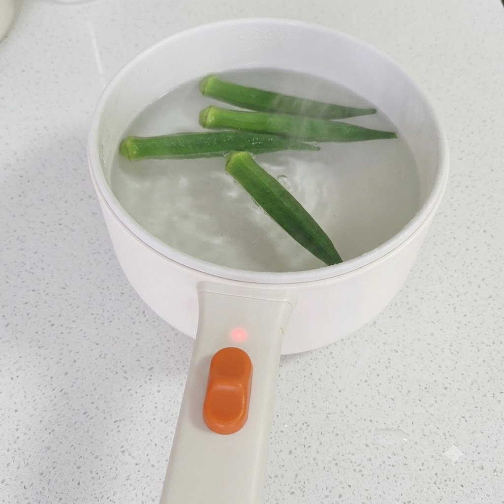 Palillos levantando okra verde cocida de una olla con agua hirviendo a fuego lento.