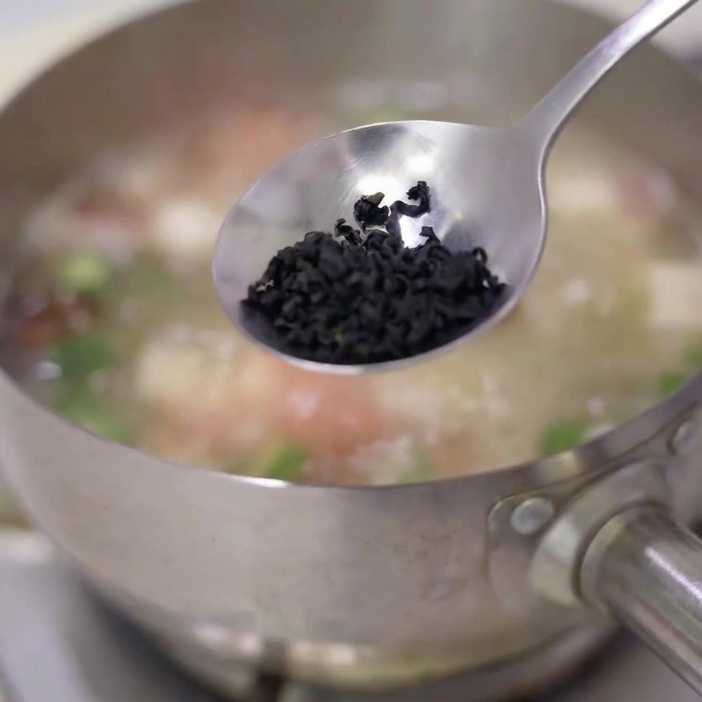 Una cucharada de hojuelas de alga deshidratada cayendo en una olla de acero inoxidable con sopa a fuego lento.