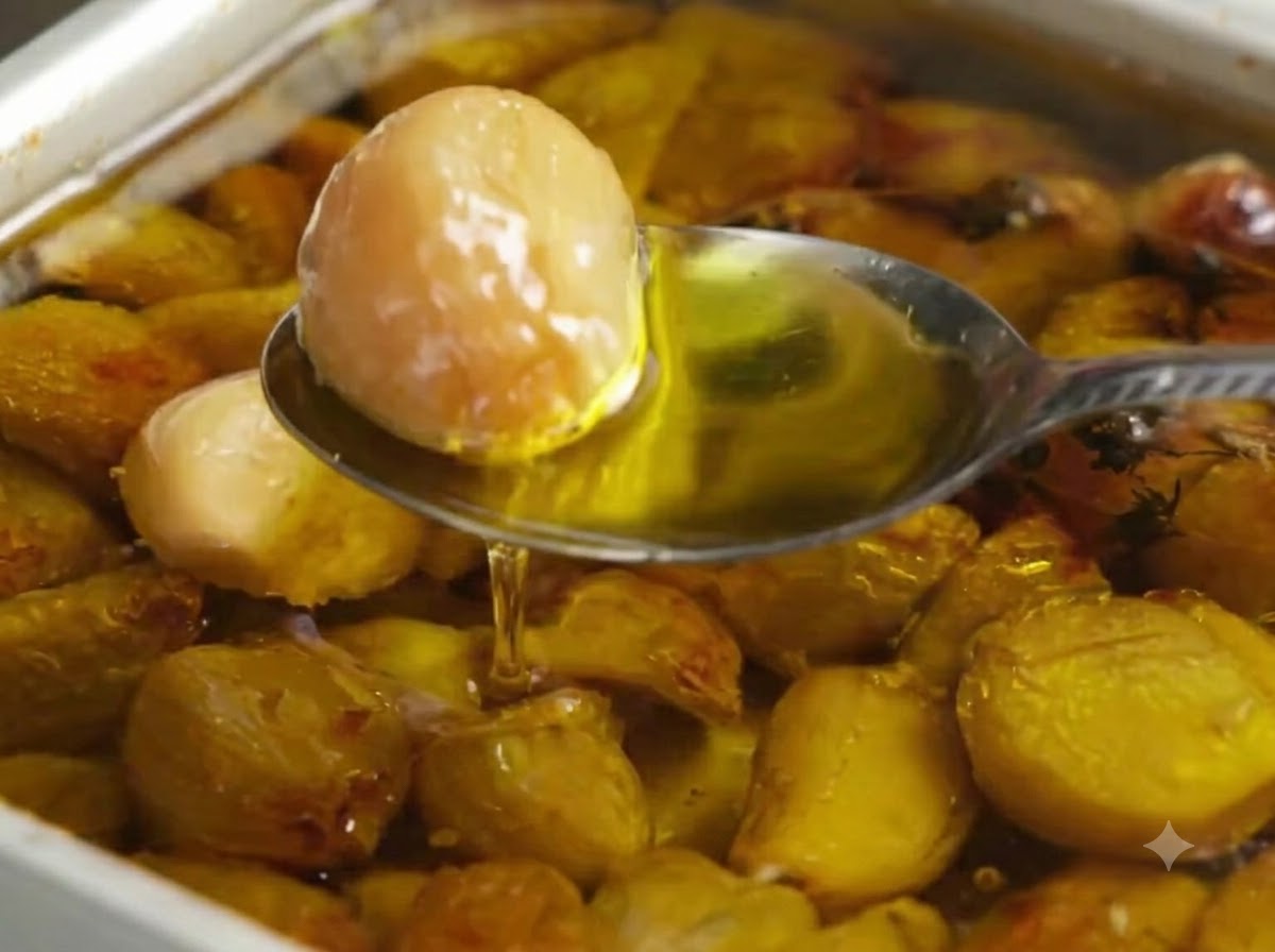Una cuchara recogiendo un diente de ajo asado dorado de una bandeja de horno llena de aceite de oliva y dientes de ajo.