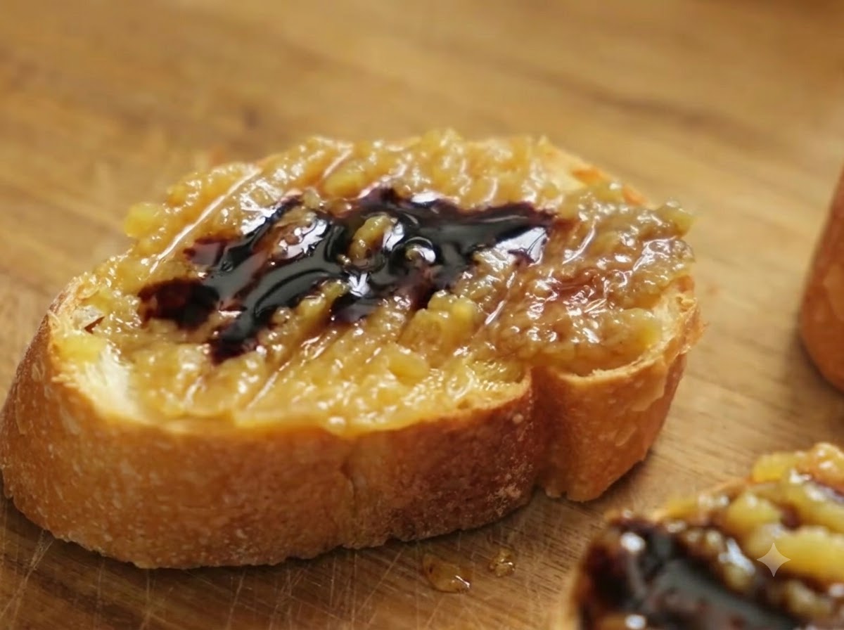 Una vista de cerca de una rebanada de baguette cubierta con pasta de confit de ajo y vinagre balsámico, con sal marina gruesa siendo espolvoreada sobre ella.