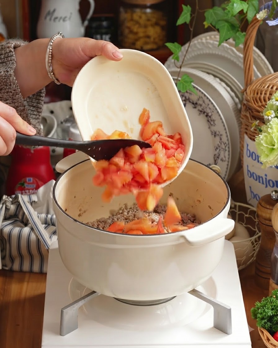 Tomates pelados en dados y verduras picadas siendo añadidos a la olla sobre la carne.