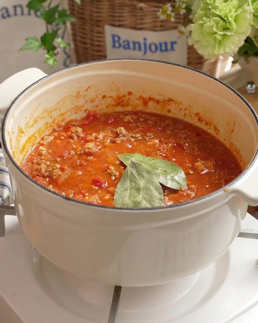 Hojas de laurel y agua fresca añadidas a la salsa de carne de tomate a fuego lento en una olla blanca.