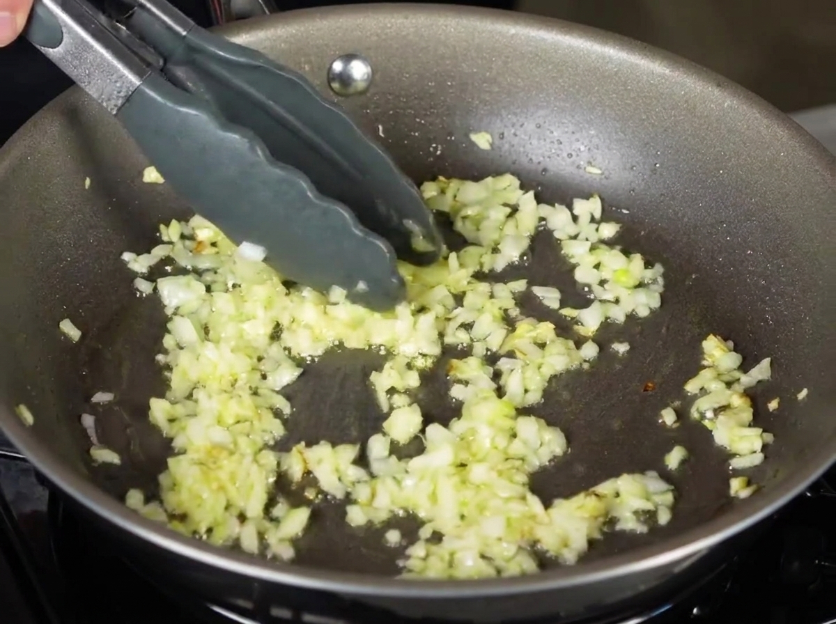 Pasta tagliatelle hervida siendo colocada con pinzas en una sartén con cebollas picadas salteadas.