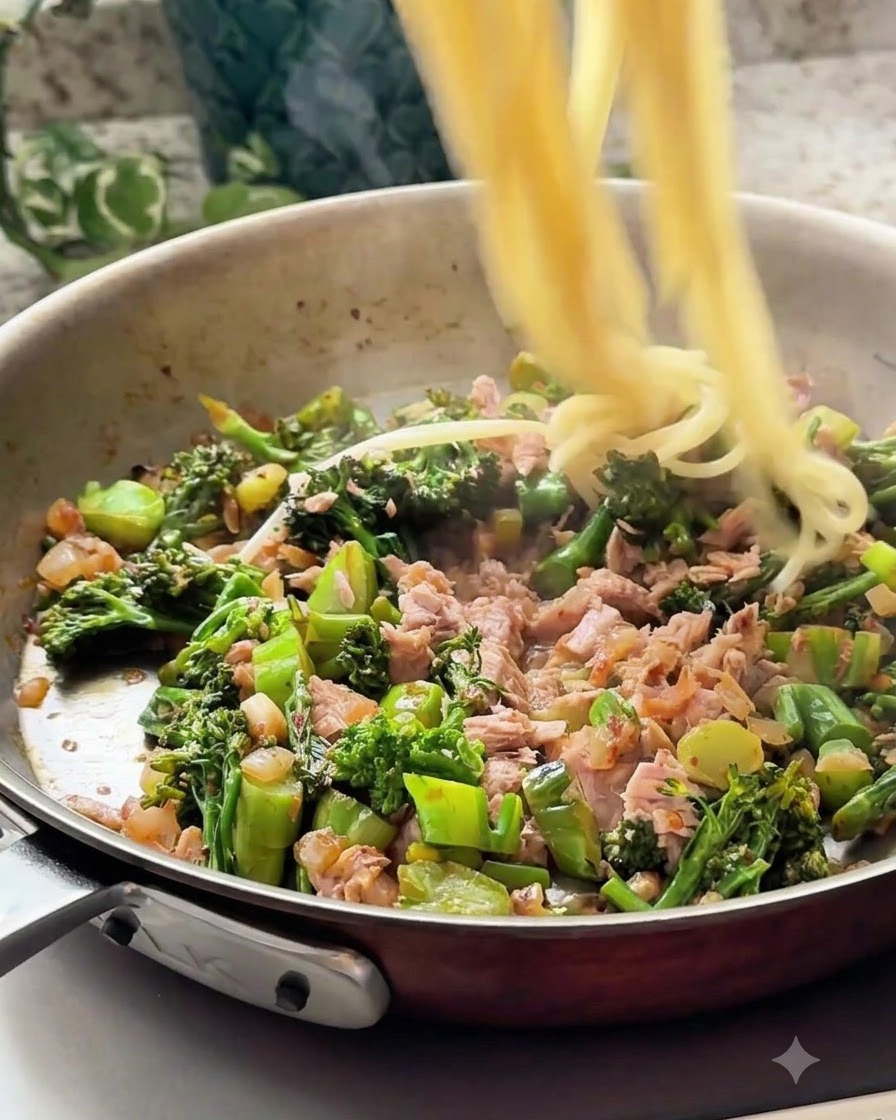 Hebras de espagueti cocido siendo incorporadas a la sartén con la mezcla de atún y verduras.