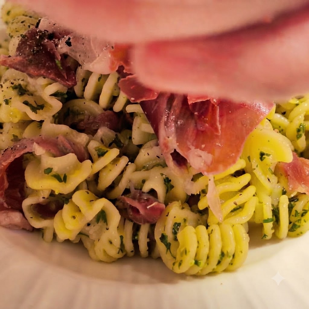 Pasta espiral recién cocida siendo transferida al bol con la salsa preparada de mantequilla y ajo, decorada con hierbas frescas y jamón.