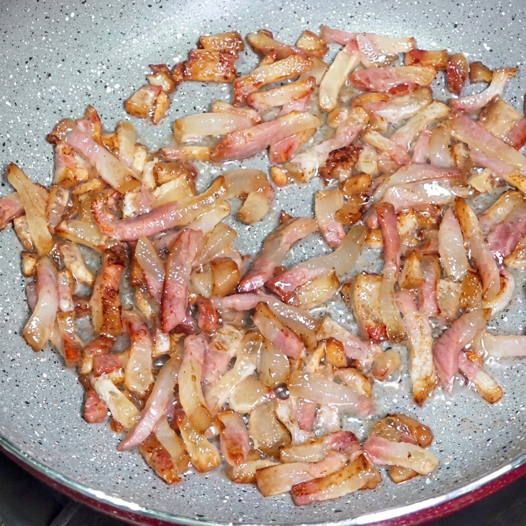 Tiras de guanciale friéndose en una sartén, liberando grasas transparentes y desarrollando bordes crujientes dorados.
