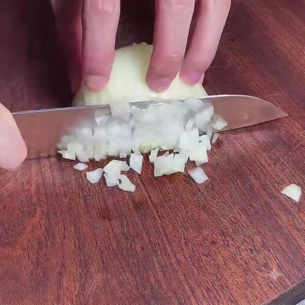 Manos sosteniendo un cuchillo para picar finamente una cebolla en una tabla de cortar de madera.