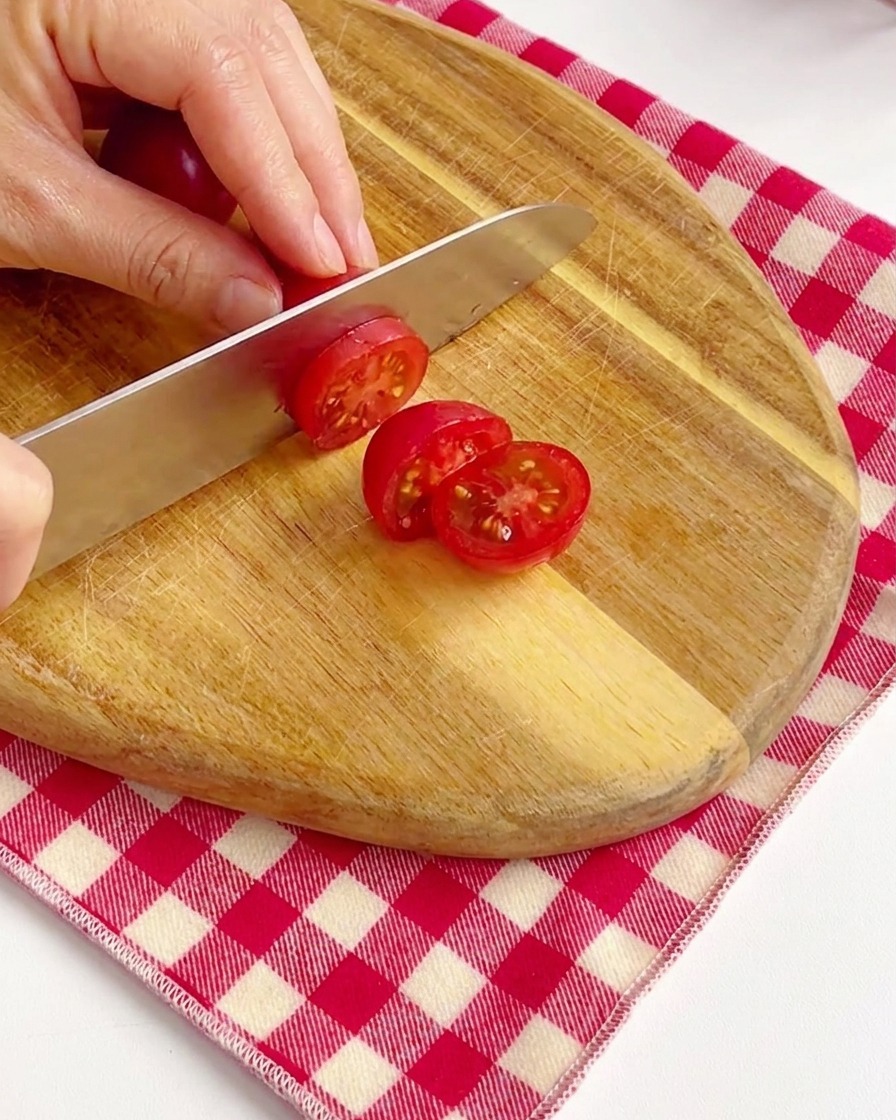 Manos cortando tomates cherry de color rojo brillante por la mitad en una tabla de cortar de madera.