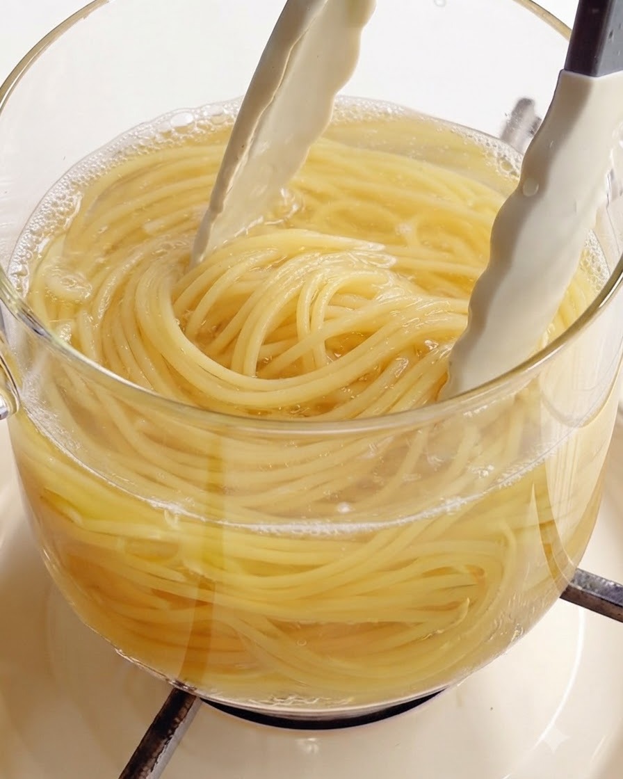 Usando pinzas de cocina blancas para sacar espagueti perfectamente cocido de una olla de vidrio transparente con agua hirviendo.