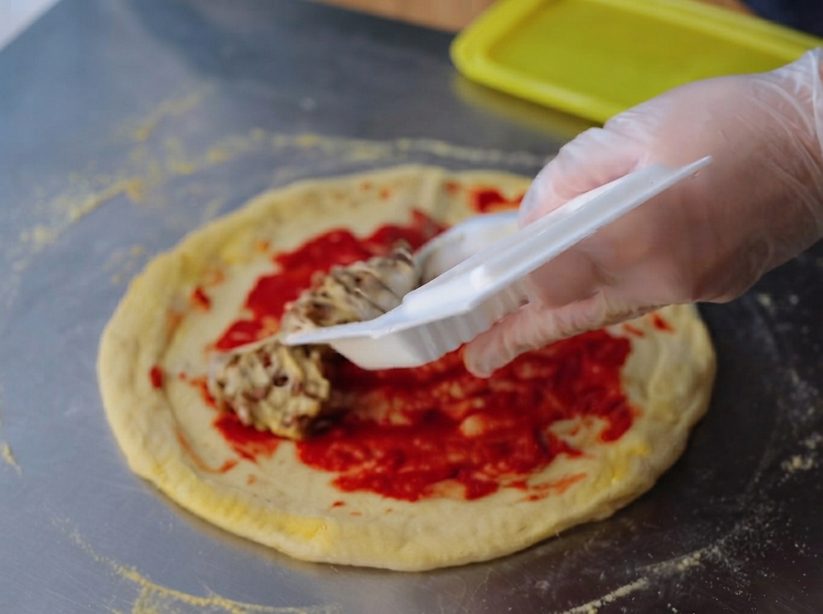 Una mano con guante extendiendo una mezcla viscosa de natto uniformemente sobre la pasta de tomate roja en la base de la pizza.