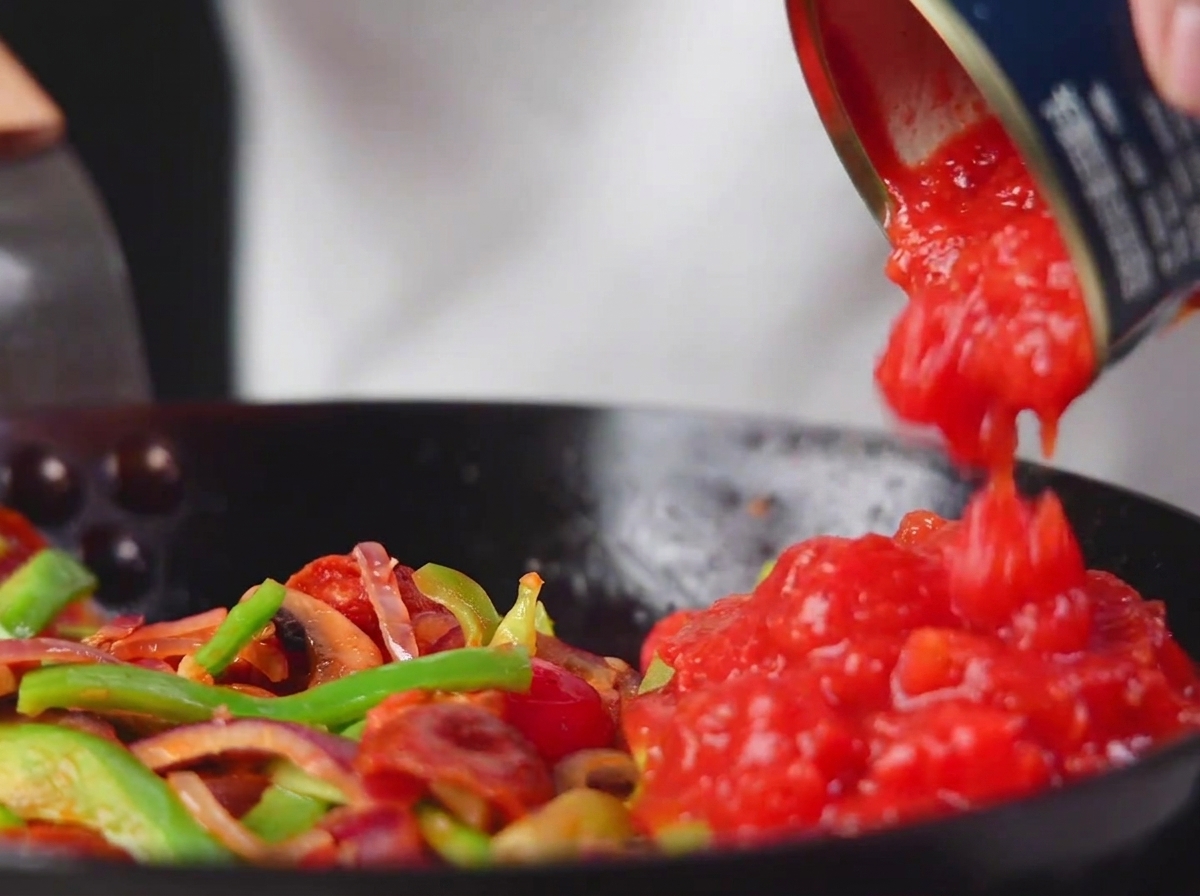 Tomates troceados enlatados siendo vertidos en la sartén con verduras y salami.