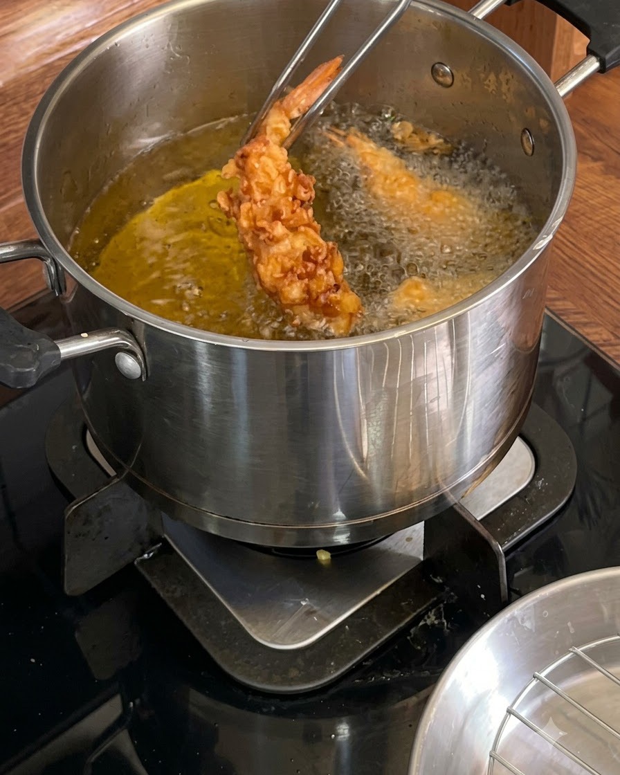 Pinzas de metal sosteniendo un camarón frito dorado y crujiente sobre una olla de aceite caliente.