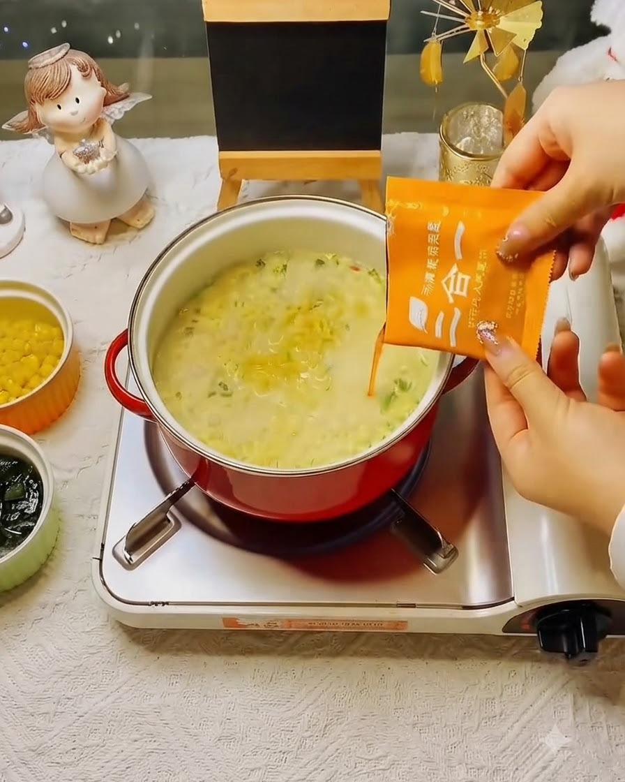 Un sobre de sopa de condimento naranja siendo vertido en la olla de fideos a fuego lento.