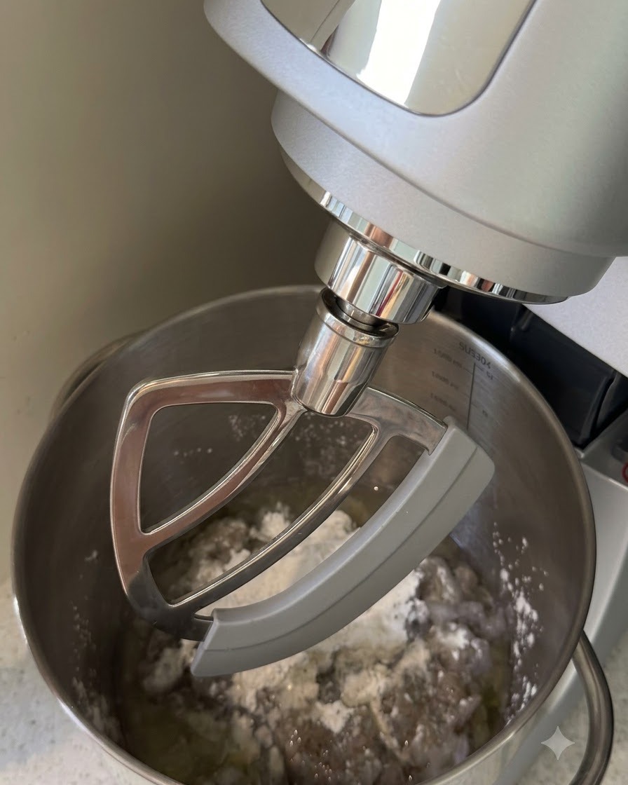Una batidora de pie con accesorio de pala mezclando camarón picado, almidón y líquidos en un bol de acero inoxidable.