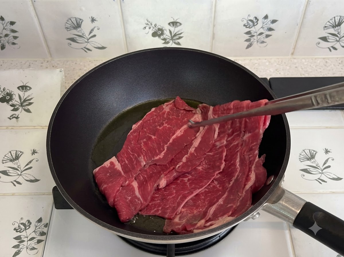 Carne de res grasa en rodajas finas salteándose en una sartén oscura con pinzas metálicas.