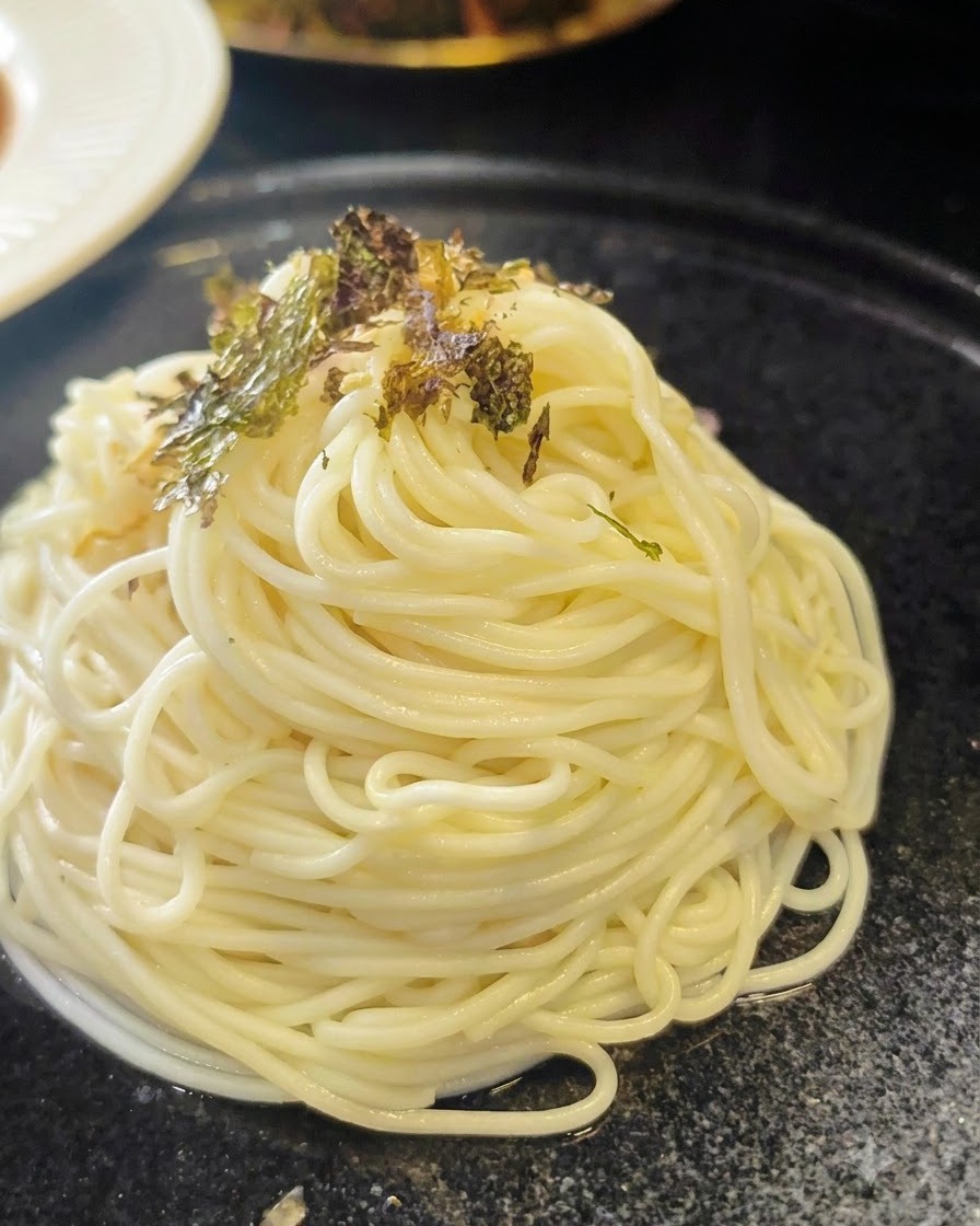 Copos de algas secas trituradas espolvoreándose sobre un montículo ordenado de fideos ramen cocidos en un plato oscuro.
