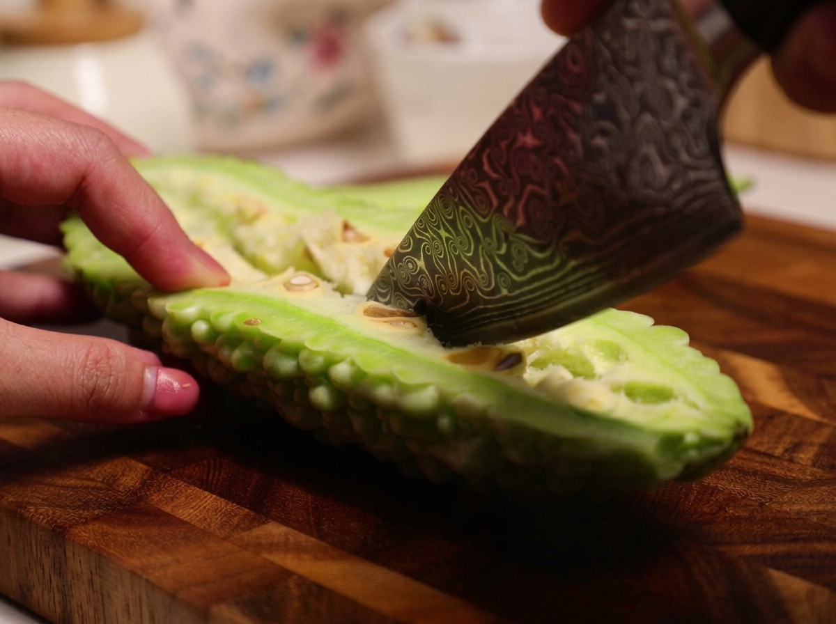 Un cuchillo siendo utilizado para raspar la pulpa blanca esponjosa y las semillas del interior de un melón amargo crudo partido por la mitad.