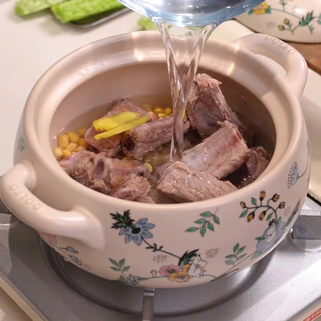Agua siendo vertida desde una jarra en una olla de guiso de cerámica decorada llena de costillas de cerdo y soja.