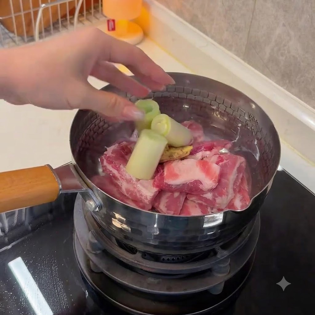 Una mano colocando trozos grandes de cebolla verde y jengibre en rodajas en una olla que contiene costillas de cerdo y agua.