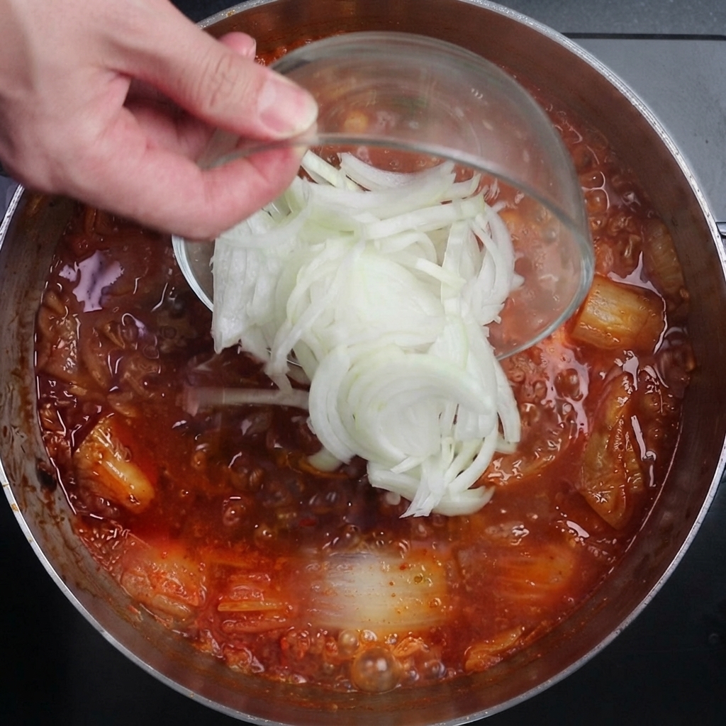 Cebollas blancas en rodajas siendo vertidas desde un tazón de vidrio en una olla con caldo de kimchi rojo, picante y hirviendo vigorosamente.
