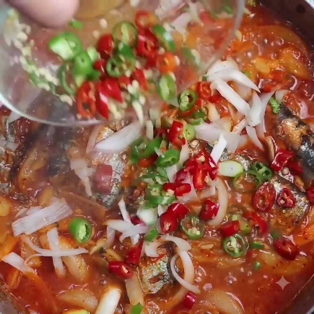 Chiles rojos y verdes en rodajas siendo vertidos desde un pequeño tazón transparente en una olla humeante de sardinas estofadas.