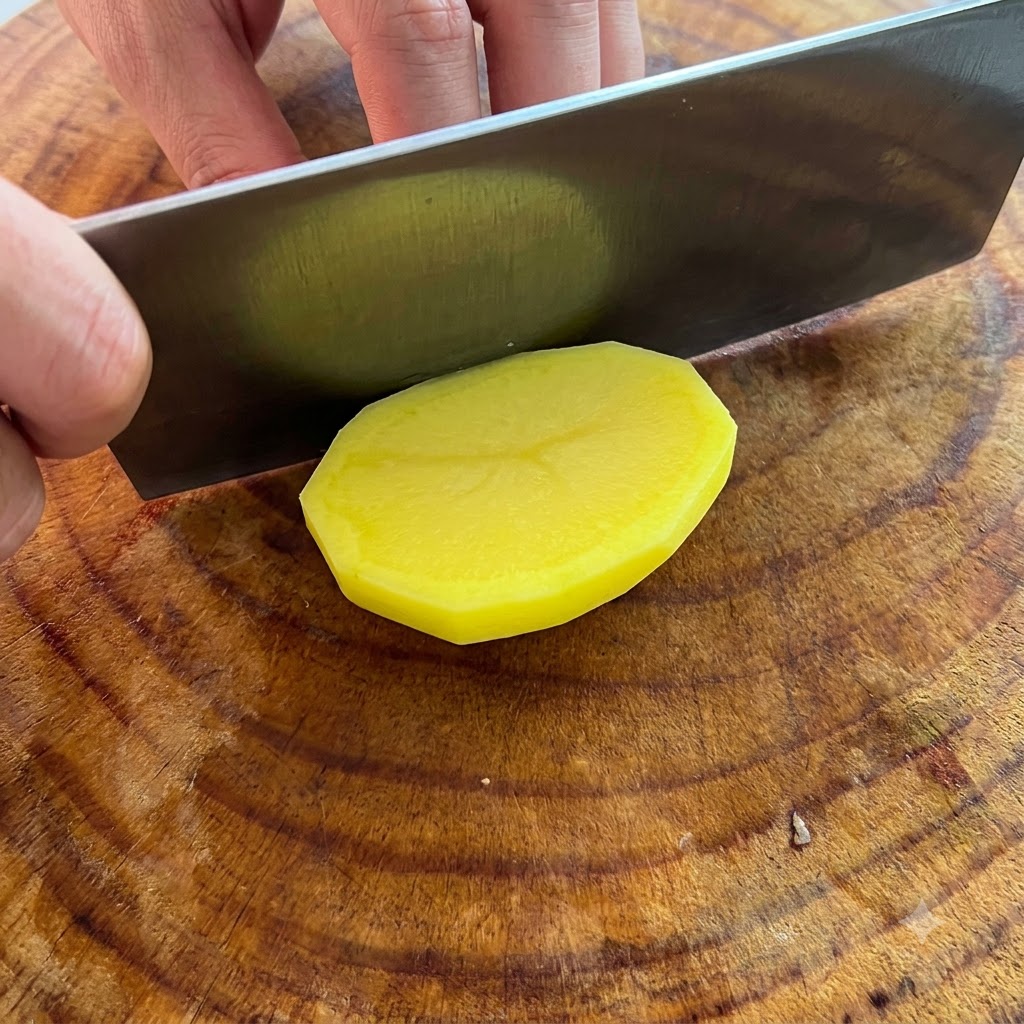 Un cuchillo cortando una papa pelada de color amarillo brillante en rodajas gruesas sobre una tabla de cortar de madera.
