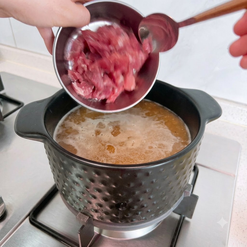 Trozos de cerdo crudo siendo vertidos desde un cuenco de metal en una olla de sopa hirviendo.