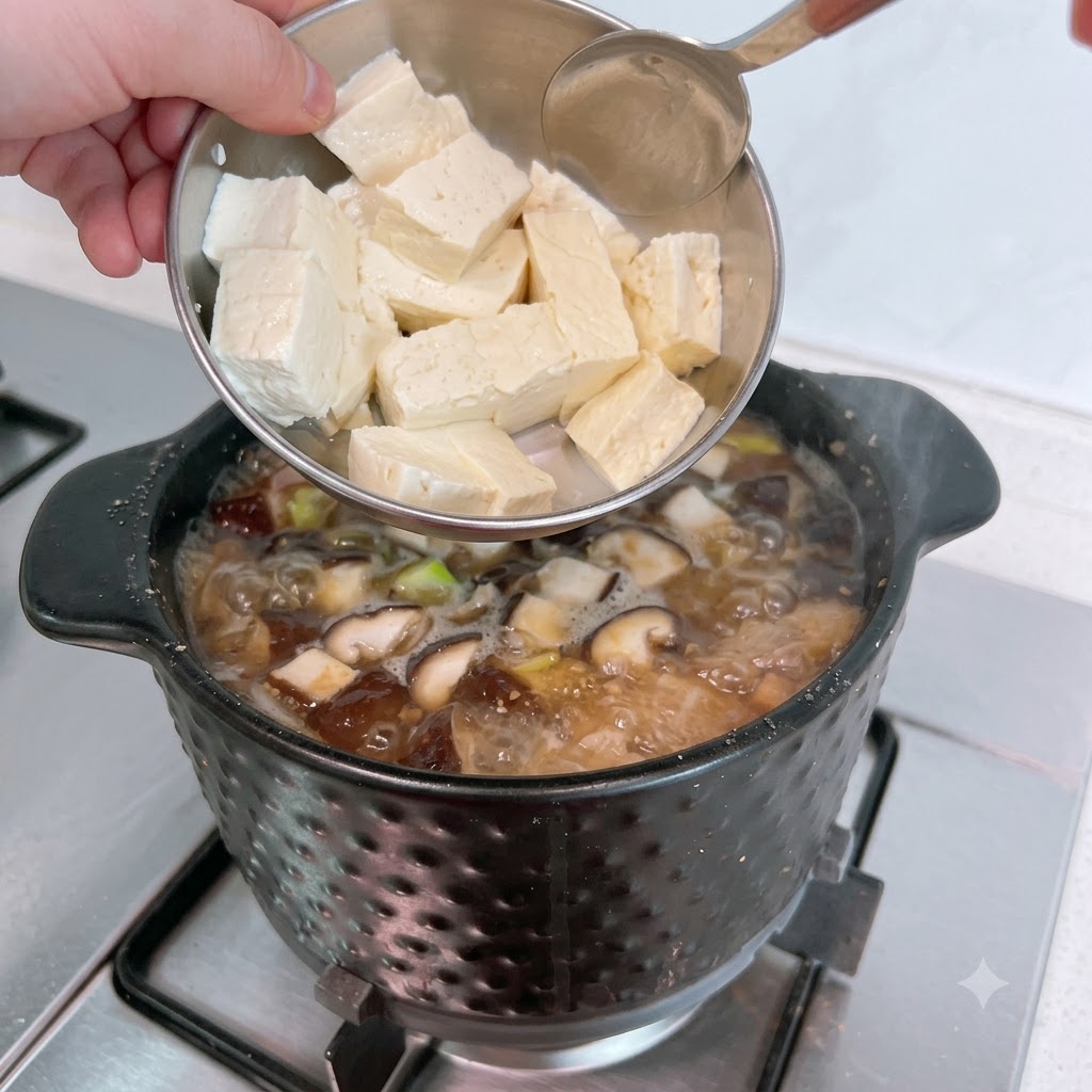 Tofu blanco en rodajas siendo añadido desde un cuenco de metal en el estofado burbujeante.