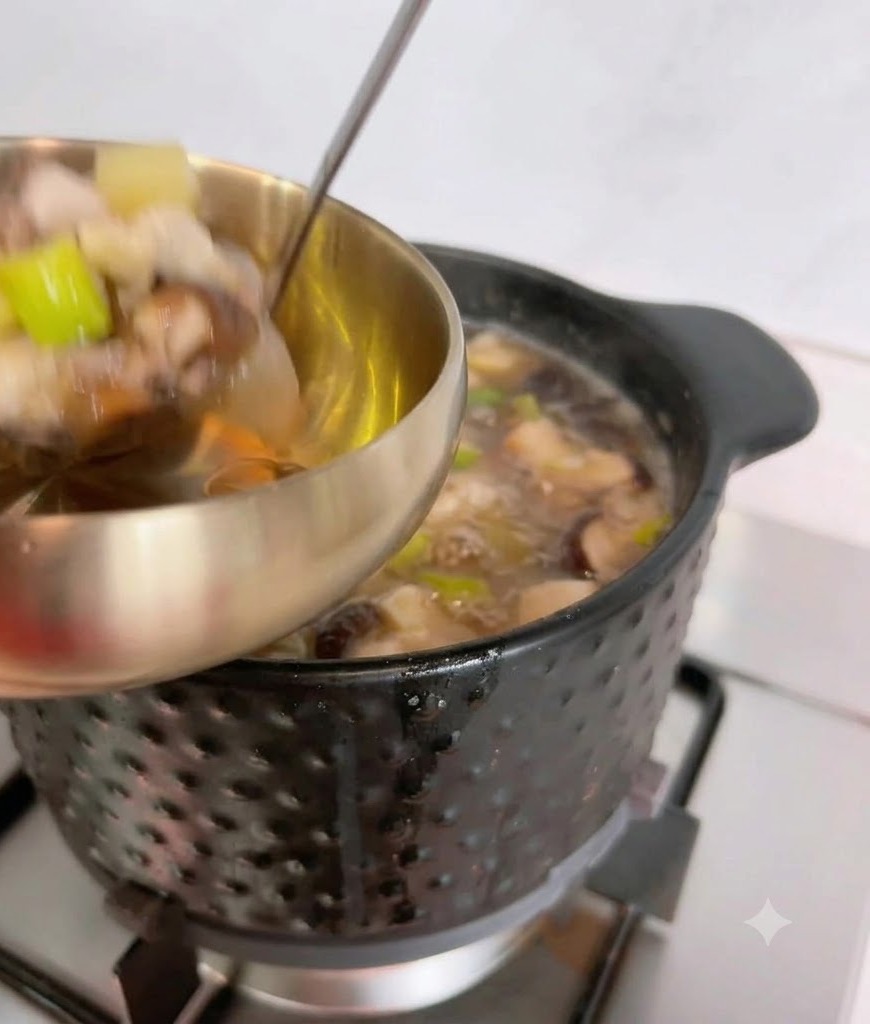 Un cucharón sirviendo sopa de pasta de soja caliente e ingredientes desde una olla negra a un cuenco de servir metálico.