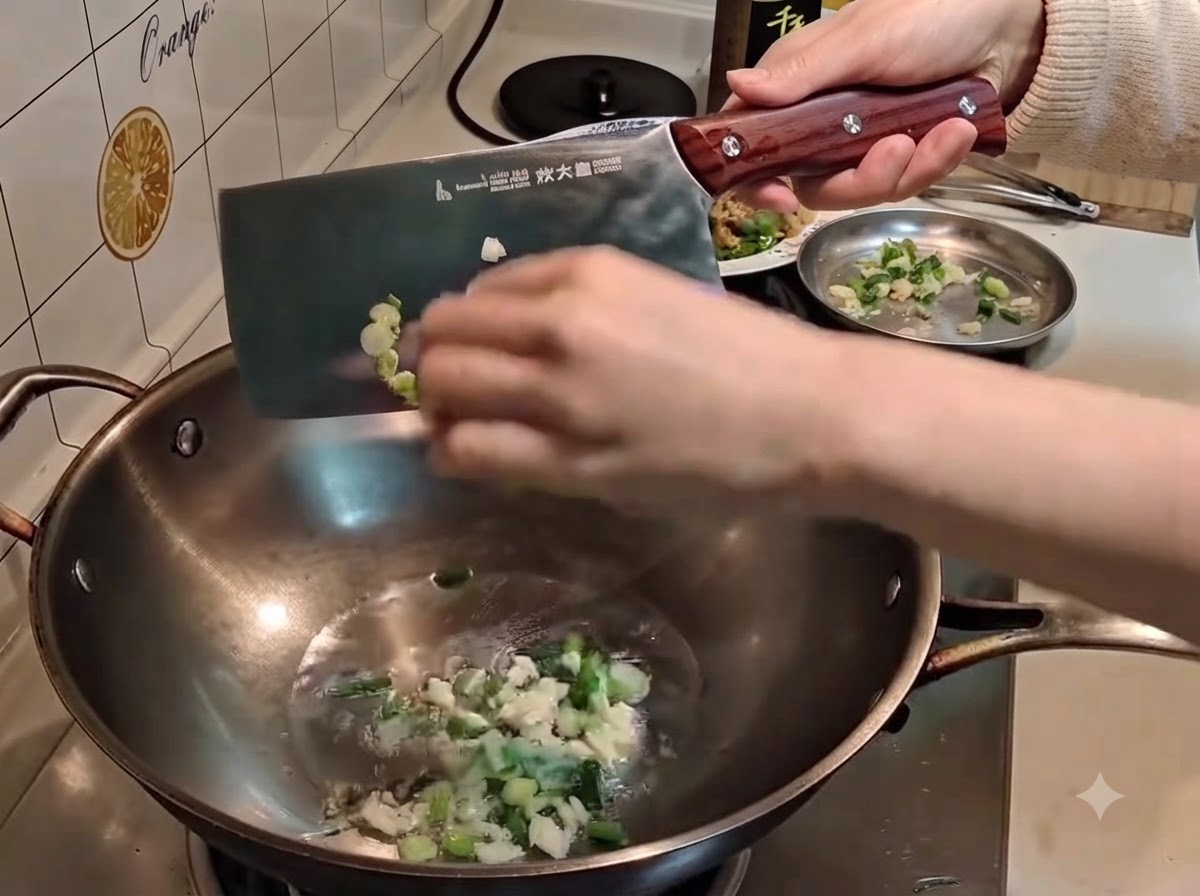 Una mano usando un cuchillo de carnicero grande de metal para empujar las cebolletas y el ajo picados en el aceite caliente de un wok.