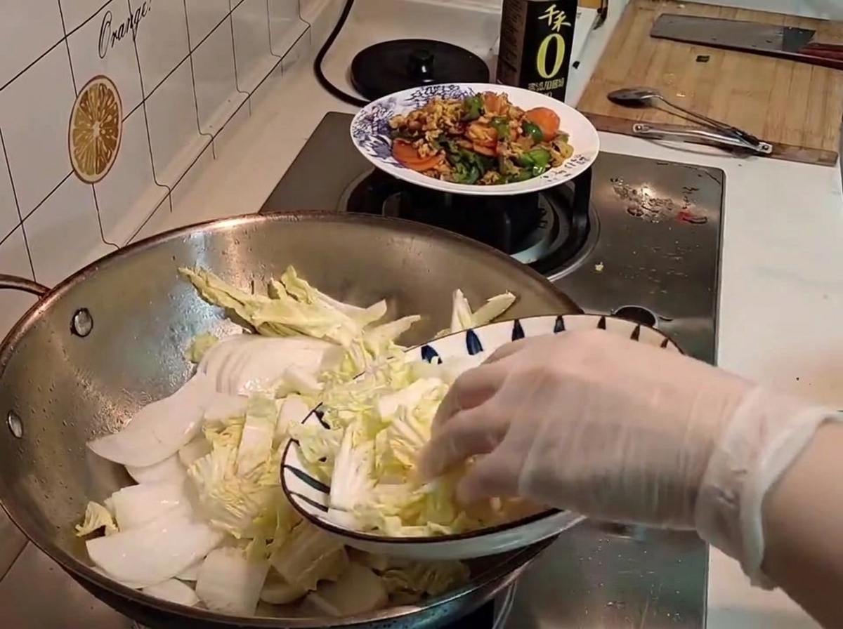 Una mano con guante transfiriendo col china cruda recién cortada desde un plato estampado a un wok.