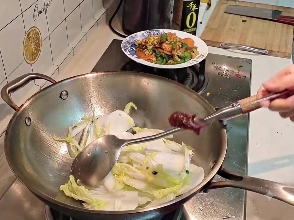 Una mano sosteniendo una cuchara de metal cargada con pasta de chile coreano rojo oscuro sobre un wok lleno de col y rábano.