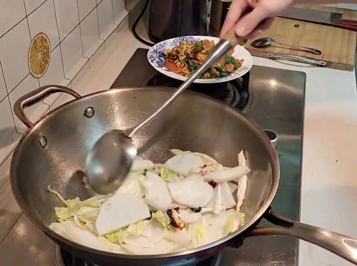 Una espátula de metal mezclando trozos de col china y rábano blanco con pasta de chile rojo dentro de un wok caliente.
